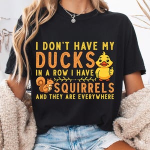 May include: A black short-sleeved t-shirt featuring yellow and orange text that reads I DON'T HAVE MY DUCKS IN A ROW I HAVE SQUIRRELS AND THEY ARE EVERYWHERE. The design includes a yellow cartoon duck giving a thumbs up and a brown squirrel.