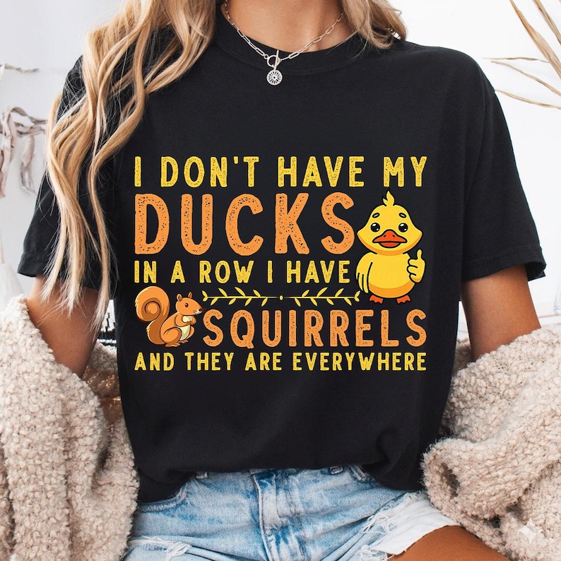 May include: A black short-sleeved t-shirt featuring yellow and orange text that reads I DON'T HAVE MY DUCKS IN A ROW I HAVE SQUIRRELS AND THEY ARE EVERYWHERE. The design includes a yellow cartoon duck giving a thumbs up and a brown squirrel.