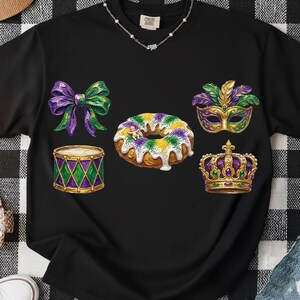 Puede incluir: Camiseta negra con gráficos coloridos con temática de Mardi Gras. El diseño incluye un lazo morado y verde, un tambor, una rosquilla, una máscara y una corona. La camiseta tiene cuello redondo y mangas cortas.