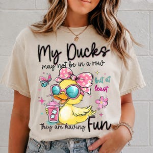 May include: A beige t-shirt featuring a graphic of a yellow duck wearing pink sunglasses and a floral bow, holding a pink drink. The text reads: My Ducks may not be in a row but at least they are having Fun, surrounded by butterfly and flower icons.
