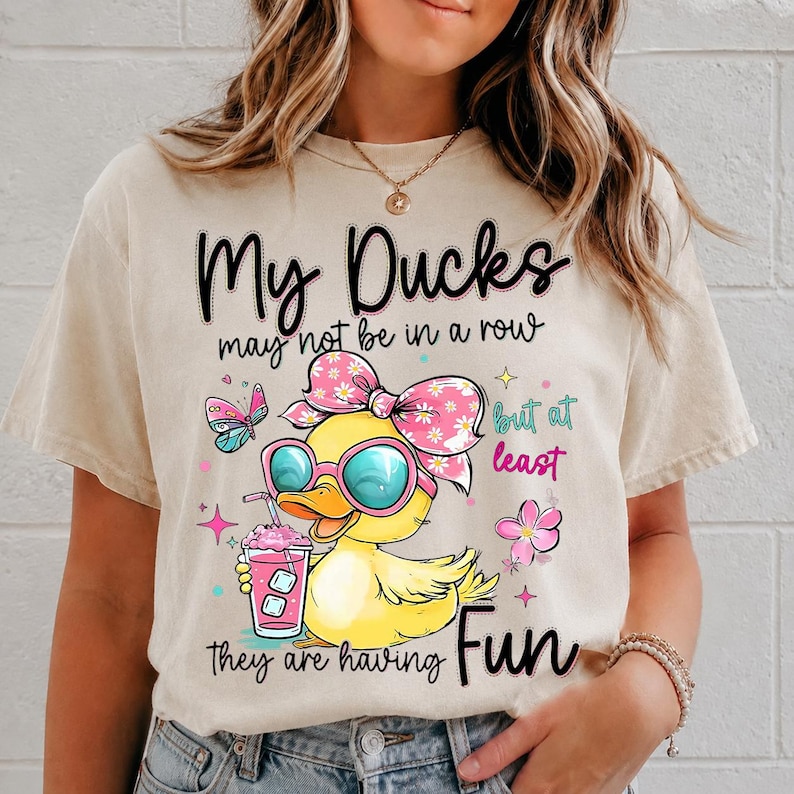 May include: A beige t-shirt featuring a graphic of a yellow duck wearing pink sunglasses and a floral bow, holding a pink drink. The text reads: My Ducks may not be in a row but at least they are having Fun, surrounded by butterfly and flower icons.