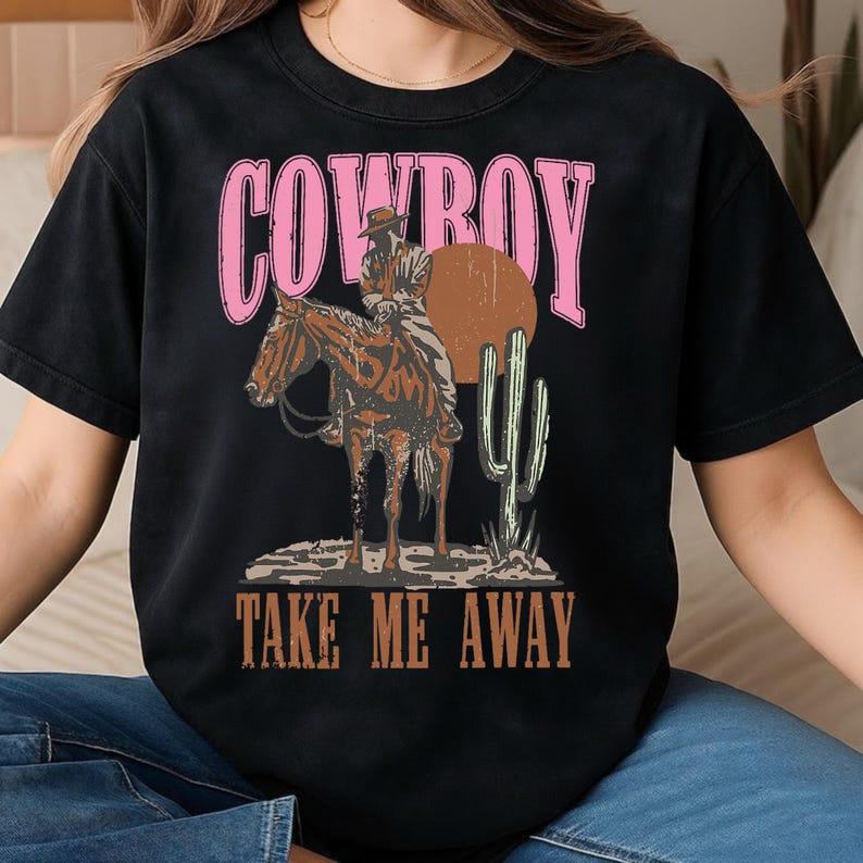 May include: Black t-shirt with a graphic design featuring a cowboy on a horse, a brown sun, and a cactus. The word "COWBOY" is in pink, and "TAKE ME AWAY" is in brown.