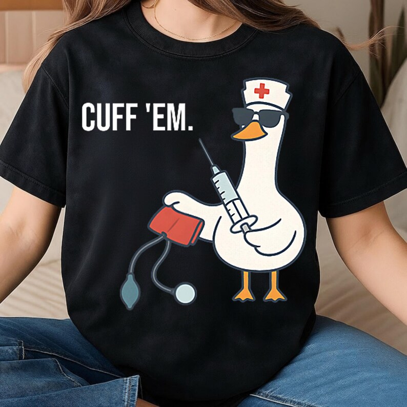Puede incluir: Camiseta negra con el texto "CUFF 'EM." sobre un dibujo animado de un ganso con gafas de sol y un gorro de enfermera, sosteniendo una jeringa y un manguito de presi&oacute;n arterial. El ganso tiene patas naranjas y un pico amarillo.