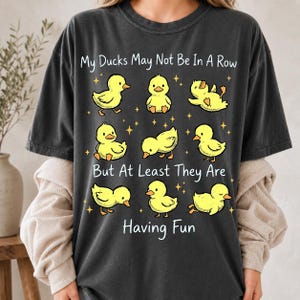 May include: A charcoal gray short-sleeve t-shirt featuring a graphic of nine yellow ducklings in various poses. The text reads, My Ducks May Not Be In A Row But At Least They Are Having Fun, surrounded by small yellow stars on the front of the shirt.