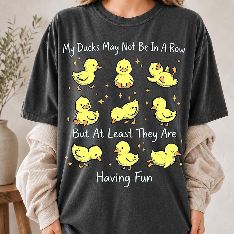 May include: A charcoal gray short-sleeve t-shirt featuring a graphic of nine yellow ducklings in various poses. The text reads, My Ducks May Not Be In A Row But At Least They Are Having Fun, surrounded by small yellow stars on the front of the shirt.