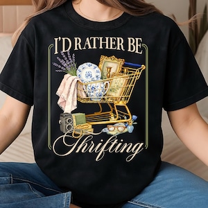 May include: Black t-shirt with a graphic of a gold shopping cart filled with vintage items, including a teacup, framed pictures, and lavender. The text reads "I'd Rather Be Thrifting" in a decorative font.