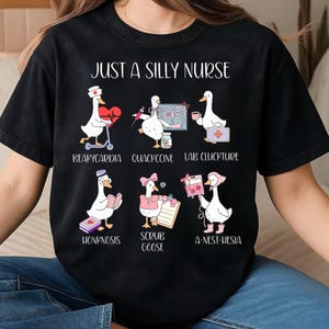 Puede incluir: Camiseta negra con el texto "JUST A SILLY NURSE" e ilustraciones de gansos en diversas situaciones m&eacute;dicas. Los gansos se representan con herramientas y accesorios m&eacute;dicos, con t&eacute;rminos m&eacute;dicos humor&iacute;sticos.