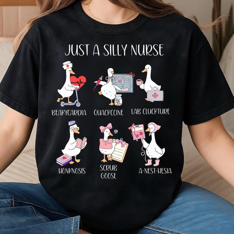 Puede incluir: Camiseta negra con el texto "JUST A SILLY NURSE" e ilustraciones de gansos en diversas situaciones m&eacute;dicas. Los gansos se representan con herramientas y accesorios m&eacute;dicos, con t&eacute;rminos m&eacute;dicos humor&iacute;sticos.