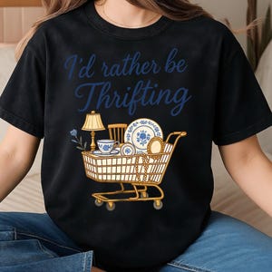 May include: Black t-shirt with the phrase "I'd rather be Thrifting" in blue script. A shopping cart graphic is filled with vintage items like a lamp, teacup, plates, and jewelry, all in a blue and gold color scheme.