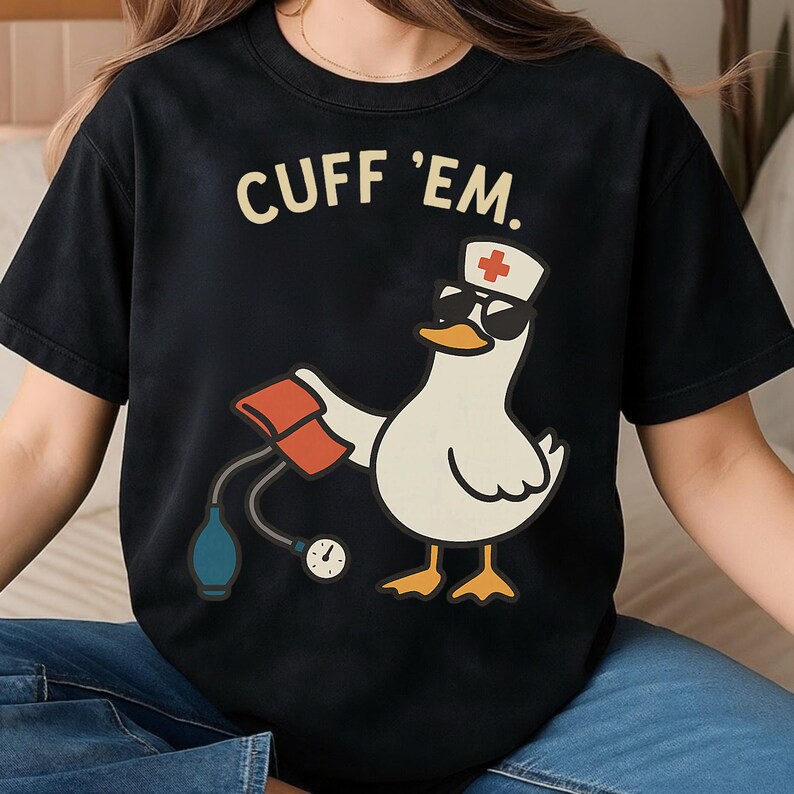 Puede incluir: Camiseta negra con un pato de dibujos animados con gafas de sol y un gorro de enfermera, sosteniendo un manguito de presi&oacute;n arterial. El texto "CUFF 'EM." est&aacute; encima del pato.