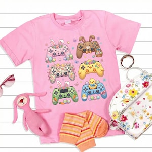Könnte beinhalten: Rosafarbenes T-Shirt mit sechs Spielcontroller-Designs, die mit Ostermotiven verziert sind. Die Controller zeigen Hasenohren, Blumen und Küken. Das Bild enthält auch einen rosa Stoffhasen, gestreifte Socken und eine Blumentasche. Ein lebendiges, auffälliges Design.
