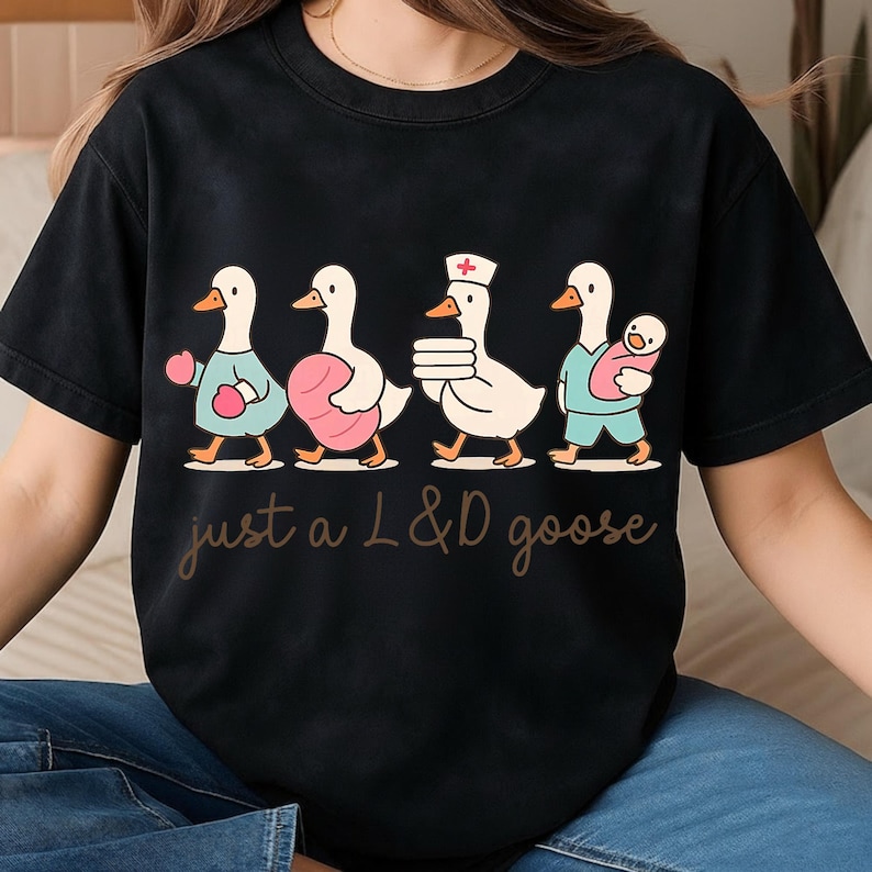 Puede incluir: Camiseta negra con una ilustraci&oacute;n caprichosa de cuatro gansos en varios roles, incluida una oca enfermera. Los gansos est&aacute;n representados con trajes azul claro, rosa y blanco. El texto "just a L&D goose" est&aacute; escrito en cursiva.