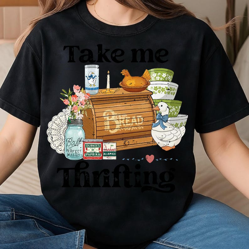 May include: Black t-shirt with the text "Take me Thrifting" above a graphic of a bread box, bowls, a duck, and other vintage kitchen items. The design includes a mason jar, spices, and a candle.