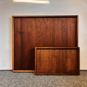 May include: Two rectangular wooden trays in a deep, dark brown hue. The larger tray is behind a smaller one, both displaying the natural wood grain. The trays have a simple, clean design, suitable for serving or display, with a classic aesthetic.