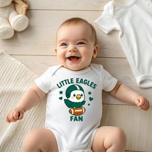 May include: A smiling baby wearing a white onesie with the text "LITTLE EAGLES FAN" and a cartoon eagle wearing a football helmet. The onesie has a football graphic and green accents.