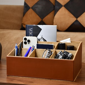 Pode incluir: Um organizador de mesa de couro marrom com vários compartimentos, contendo canetas, um smartphone, óculos e um livro. O organizador está em uma mesa de madeira, com almofadas decorativas ao fundo.