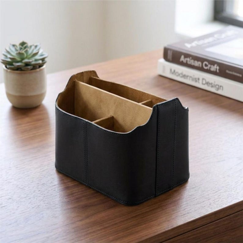 May include: A black leather desk organizer with multiple compartments. The organizer has a rectangular shape with curved edges and a light brown interior. A small succulent plant in a beige pot and two books are in the background.