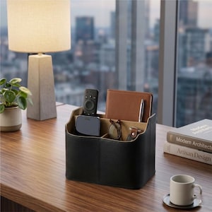May include: A black leather desk organizer with multiple compartments, holding a remote, a small device, a notebook, glasses, and a pen. The organizer sits on a wooden desk, with a lamp and a plant in the background.
