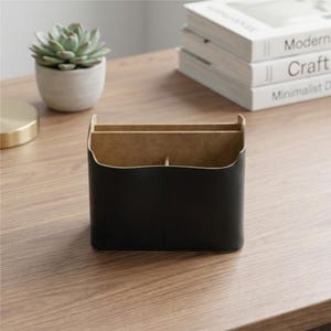 May include: A black leather desk organizer with two compartments and a central divider. The organizer has a light brown interior and is sitting on a wooden surface. A small potted succulent and a stack of books are in the background.