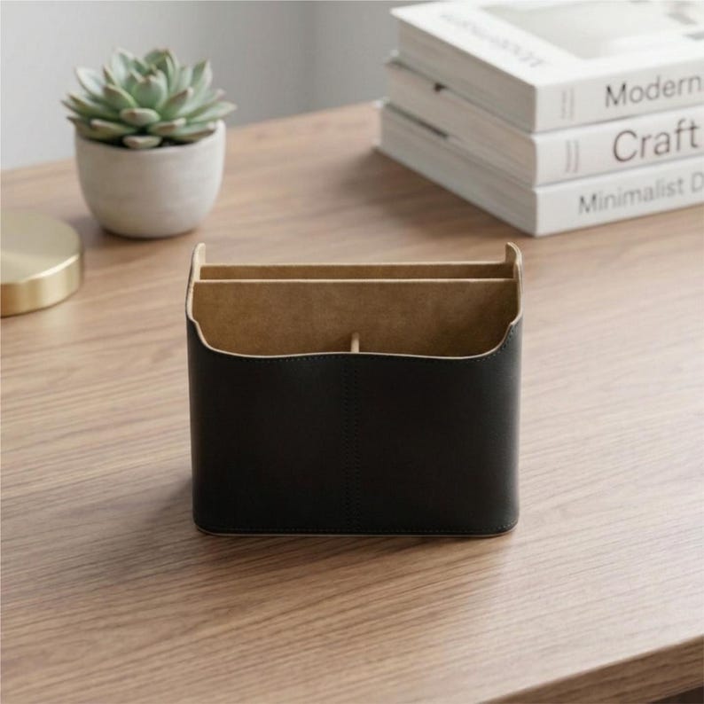 May include: A black leather desk organizer with two compartments and a central divider. The organizer has a light brown interior and is sitting on a wooden surface. A small potted succulent and a stack of books are in the background.