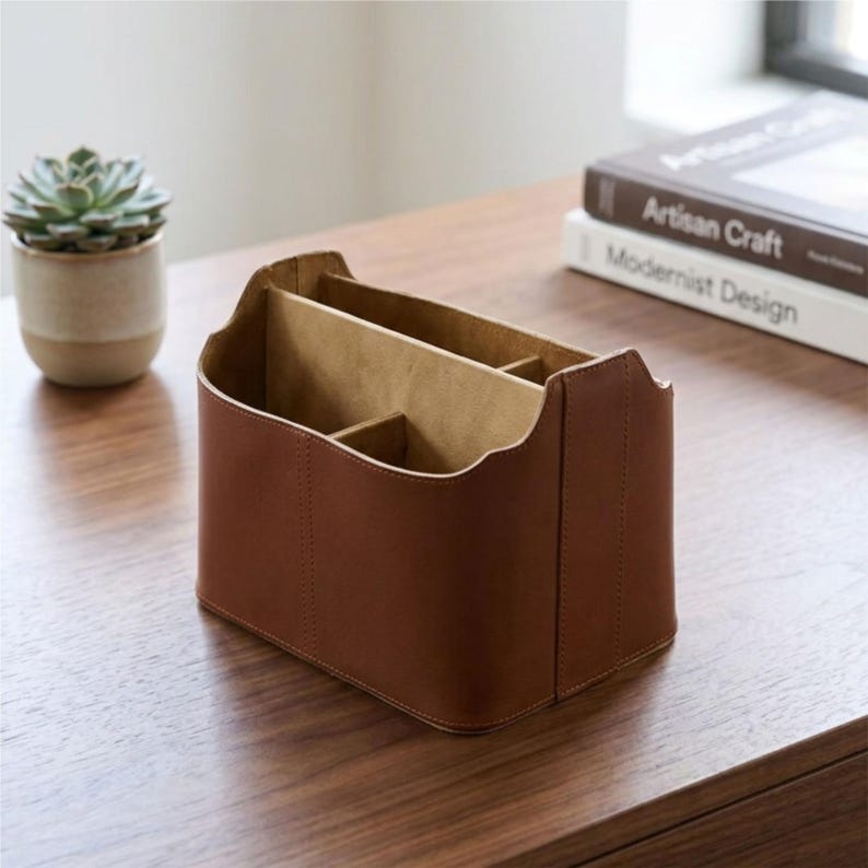 May include: A brown leather desk tidy with several compartments. The rectangular organiser has a curved top edge and is on a wooden surface. A small potted succulent and two books are also visible.