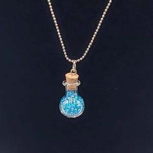 May include: A silver-colored necklace with a small glass bottle pendant. The bottle is filled with small blue beads and sealed with a cork. The background is a solid black.