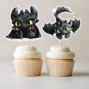 Op de afbeelding: Twee cupcakes met zwarte draaktoppers met groene ogen en roze accenten. De cupcakes hebben witte frosting en staan tegen een neutrale achtergrond. De toppers zijn bevestigd aan houten stokjes.