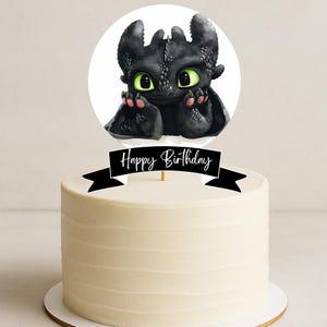 Puede incluir: Una tarta blanca glaseada con una pancarta negra de "Feliz Cumpleaños" y un adorno circular con un dragón negro con ojos verdes. El dragón está en una pose linda, agregando un toque caprichoso a la tarta.