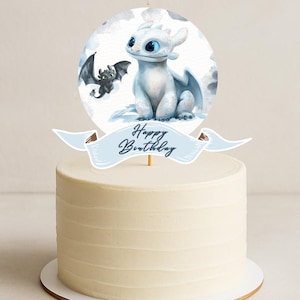 May include: A white frosted cake topped with a round image of a white dragon and a black dragon. A light blue banner with the words "Happy Birthday" is in front of the image. The cake is on a white cake stand.