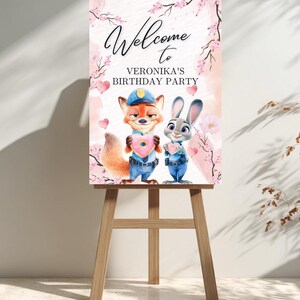 Puede incluir: Un letrero de bienvenida para la fiesta de cumplea&ntilde;os de Ver&oacute;nica, con los personajes de dibujos animados Nick Wilde y Judy Hopps sosteniendo donuts en forma de coraz&oacute;n. El letrero est&aacute; decorado con flores de cerezo rosas y corazones, exhibido en un caballete de madera.
