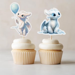 Op de afbeelding: Twee cupcakes met witte glazuur en draak-thema toppers. Eén draak houdt een blauwe ballon vast, de andere zit met gevouwen vleugels. De cupcakes hebben lichtbruine, gegolfde papieren vormpjes.