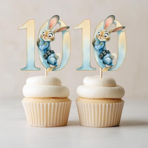 May include: Two cupcakes with white frosting, topped with a Judy Hopps character cake topper. The cake toppers feature the number 10 with a cartoon bunny character. The cupcakes sit on a light-colored surface.