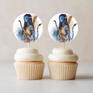 Puede incluir: Dos cupcakes con adornos circulares de papel que muestran un personaje humanoide de piel azul con cabello largo y oscuro, ojos amarillos, arco y flechas. El personaje viste una prenda color canela y tiene una cola larga sobre un fondo de acuarela.