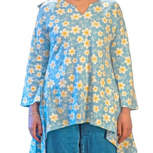 Op de afbeelding: Een lichtblauwe tuniek top met een madeliefjes bloemenpatroon. De top met lange mouwen heeft witte madeliefjes met gele centra, een V-hals en een asymmetrische zoom. De top wordt gecombineerd met een blauwe jeans.
