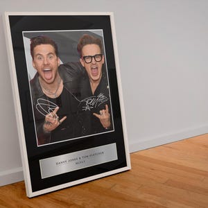 May include: Framed photograph featuring two men, Danny Jones and Tom Fletcher of McFly, with autographs. The black and white photo is set in a black frame with a white border. The frame is displayed on a wooden floor.