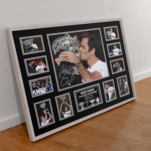 May include: Framed photo collage featuring Roger Federer kissing a trophy, surrounded by smaller images of him. The collage includes his signature and text: "2018 Australian Open Winner". The frame is white, with a black mat.