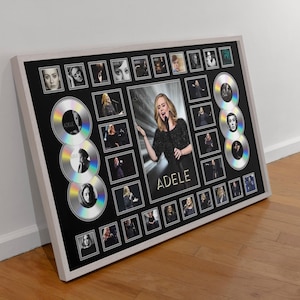 Puede incluir: Obra de arte enmarcada con una imagen central de Adele, rodeada de fotos más pequeñas y diseños de CD estilizados. El fondo negro contrasta con el marco blanco y los detalles plateados, creando una pieza visualmente impactante.