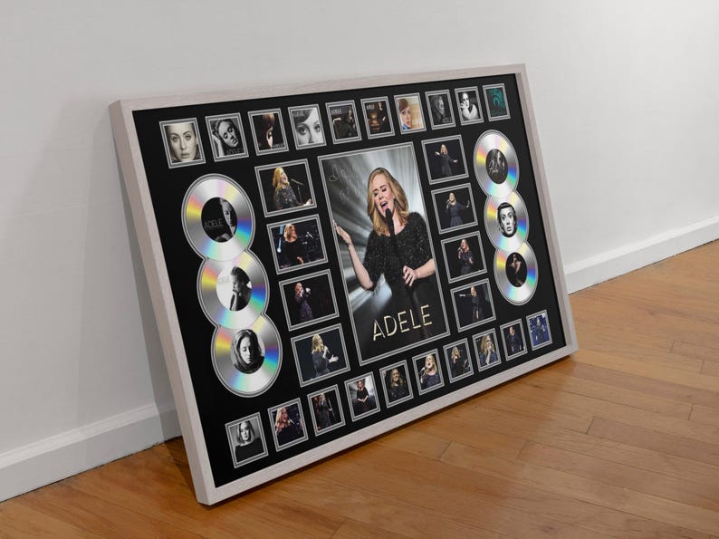 May include: Framed artwork featuring a central image of Adele, surrounded by smaller photos and stylized CD designs. The black background contrasts with the white frame and silver accents, creating a visually striking piece.