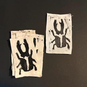 Op de afbeelding: Een verzameling kleine, rechthoekige stoffen patches met een zwart keverontwerp. De kever is afgebeeld met grote kaken en zes poten. De patches zijn bedrukt op een witte stof met een getextureerde uitstraling en zijn gerangschikt op een donkere ondergrond.