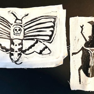 May include: Two white fabric squares with black linocut prints. One features a moth with a skull design, the other a beetle. The prints are detailed and artistic, suitable for textile art or craft projects.