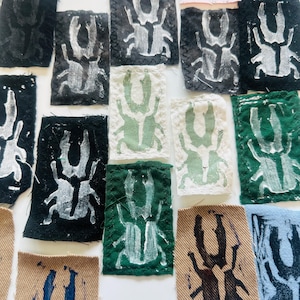 May include: A selection of fabric patches, each displaying a distinct insect design. The patches are in various colours, such as black, green, tan, and blue, with the insect illustrations in contrasting white or darker shades. The fabric pieces are hand-stitched.