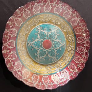May include: Decorative glass plate with a scalloped edge. The plate features concentric circles of colour: red, yellow, and teal, with silver filigree designs. A red centre dot completes the design. A beautiful piece for display.