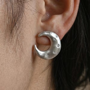 Può includere: Un orecchino a forma di mezzaluna color argento con una finitura martellata e testurizzata. La superficie della luna presenta piccoli motivi a stella incisi sparsi sulla forma curva. L'orecchino è fissato al lobo con una chiusura a perno.