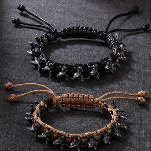 May include: Two adjustable bracelets featuring a series of metallic, silver-toned vertebrae-shaped charms woven into black and tan cord bands. Each bracelet includes a sliding knot closure with knotted ends, showcasing a detailed, segmented design.