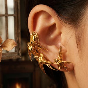 May include: Gold-colored ear cuff featuring a flock of stylized birds in flight. The jewelry piece is designed to wrap around the ear, with multiple birds extending along the ear's curve. The birds have detailed wings and bodies.