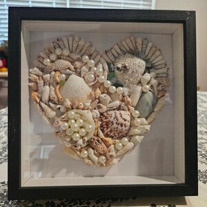 May include: A framed heart-shaped artwork composed of various seashells and faux pearls. The shells are in shades of white, beige, and brown, arranged to form the heart. The black frame provides a contrasting border, enhancing the artwork's visual appeal.
