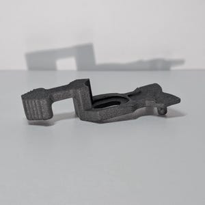 Puede incluir: Un componente mecánico de color gris oscuro, texturizado e impreso en 3D, que presenta una abertura ovalada central, un brazo rectangular con un extremo estriado y una extensión plana y cónica, con un acabado mate y estructura compleja.
