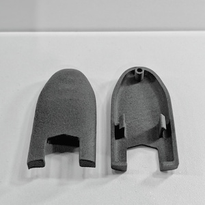 May include: Two dark gray, matte-textured, arch-shaped plastic components. One piece shows the smooth, rounded exterior surface, while the other displays the hollow interior with a small circular mounting hole and two rectangular support tabs.