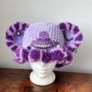 May include: A light purple crocheted bucket hat with large, fluffy purple and light purple faux fur ears. The hat features a purple embroidered patch with the text SUBTRONICS and a beaded strand with the text SPLOINKY DUB, dice, and a number 3 ball.