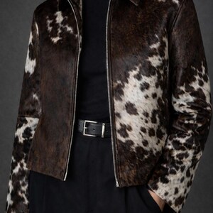 May include: A brown and white cowhide jacket with a zip-up front and a collar. The jacket is worn over a black turtleneck and paired with black pants and a black belt.